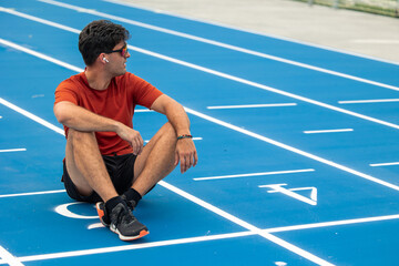 Athletic Man with Sunglasses and Wireless Earphones sitting on the Finish Line of Athletic Track