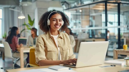 Cheerful woman wearing headphones and a light jacket smiles while working at a laptop in a sunny office