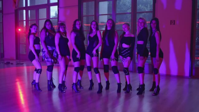 A group of female dancers in high heels poses for the camera. Nine girls stand in a row in a dance studio, posing for the camera. Purple light.