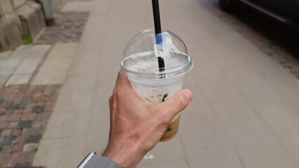 In the city, a man walks holding a beverage cup with a black straw, enjoying a refreshing drink while strolling in the urban environment