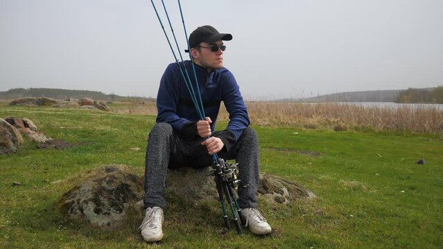 A young man, in a blue jacket, glasses, with spinning rods in his hands, sits in nature near the river, enjoying the silence and dreaming of a good catch. Slow motion.