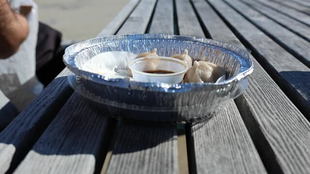 man eating chinese pork dumplings with chopsticks on a picnic table (aluminum metal takeaway container and dumpling soy sauce in plastic) asian takeout eat in park lifting hand 