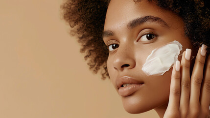 Portrait of a dark skinned woman with curly hair, applying face cream on cheek poses against a peach background.