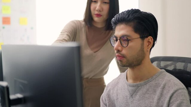 A focused male programmer, wearing glasses, is typing code on a computer, with a female engineer pointing at the monitor, guiding and collaborating with him.