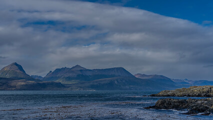 A beautiful mountain range of the Andes against a background of blue sky and clouds. In the foreground is the blue water of the ocean, the islands in the Beagle Channel. Argentina. Tierra del Fuego