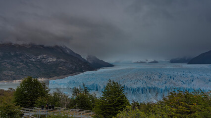 The beautiful blue glacier Perito Moreno stretches to the horizon between the mountains. A mass of ice with cracks, crevices, sharp peaks. There are tourists on the observation deck. Green vegetation 