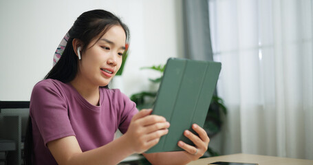 Portrait of Young asian businesswoman wear wireless headphones hold digital tablet having video call with partners or coworkers on laptop while sitting on desk at home office