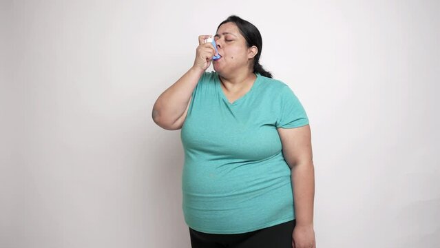 Overweight, fat indian woman having difficulty in breathing and using an asthma inhaler for relief. isolated over white background. Healthcare concept.4k