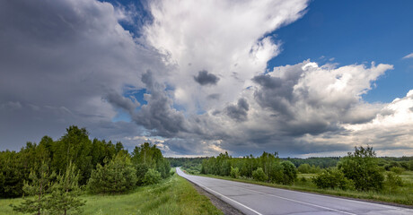 A paved road curves through a lush green forest, framed by a dramatic sky filled with fluffy white...
