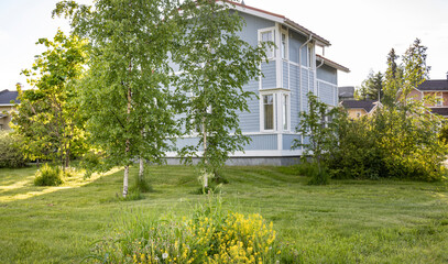 A light blue house stands on a lush green lawn, surrounded by vibrant foliage and trees. The sun casts warm shadows, creating a peaceful atmosphere.