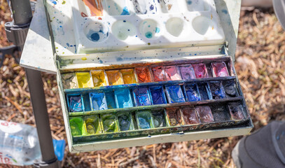 A close-up image of a watercolor palette, showcasing vibrant colors ready for use. The palette appears to be well-loved, with evidence of past artistic endeavors.