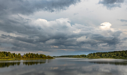 A serene lake with lush green trees on its banks reflects the gray and white clouds of a passing...