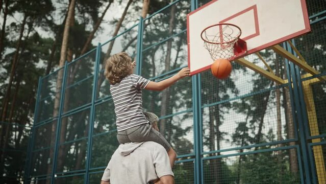 Rear of Caucasian family dad and kid boy having fun playing basketball. Happy son sitting on father's shoulders throwing a ball in hoop outdoors on court. Leisure activity. Bottom view. Sport concept