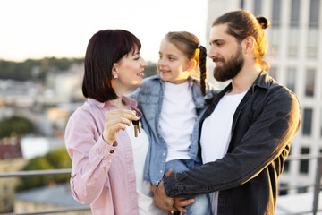 Joyful young family celebrating new home purchase on rooftop, parents holding child and showing house keys.