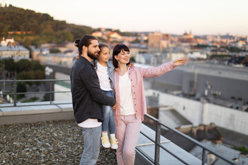 Warm and joyful moment of a happy family standing on a rooftop, enjoying city view. Man holding child, woman pointing.