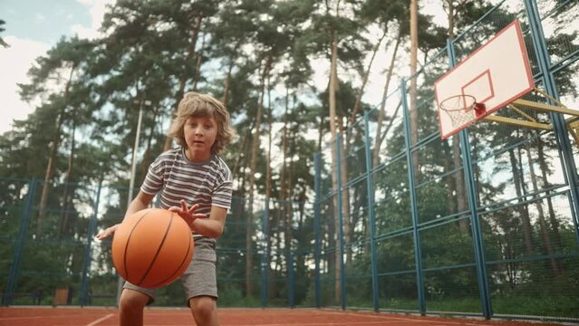 Close up of Caucasian small child playing basketball bouncing ball on court outside. Little kid dribble ball running on urban court practicing basketball moves. Sport lifestyle. Recreation, training
