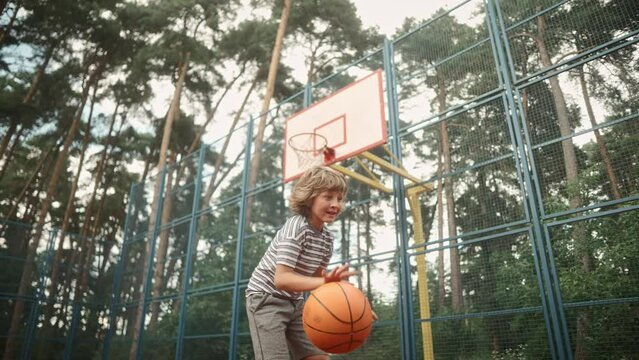 Close up of Caucasian little child playing basketball bouncing ball on court. Little kid dribble ball running on playground practicing basketball in park. Sport, recreation and leisure, bottom view