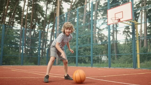 Happy positive cute teen boy bouncing ball practicing basketball moves outdoor on court. Caucasian little cheerful child playing basketball. Dribble moves. Fun. Leisure activity. Childhood concept