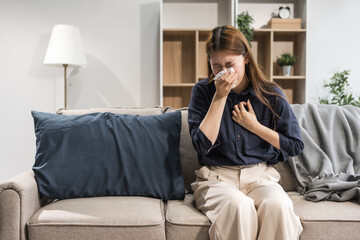 A young female Asian is sitting on a sofa, wrapped in a shawl. She is experiencing cold symptoms like a cough, sneeze, sore throat, and congestion, indicating a viral infection.