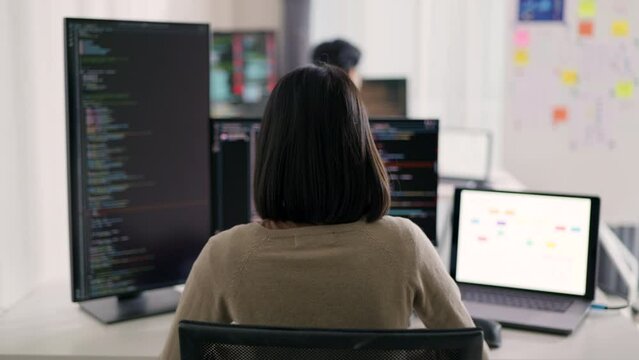 An Asian woman programmer is focused on coding at her dual-monitor workstation, surrounded by screens displaying lines of code and data.