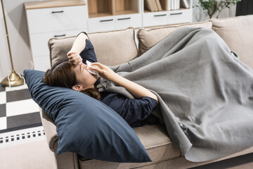 A young female Asian is resting on a sofa, suffering from the flu. She shows symptoms like fever, chills, body aches, and fatigue, requiring rest and care.