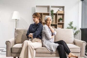 A mature mom and her young daughter, both Asian, sit on a sofa, engaged in a quarrel. Their...