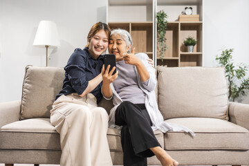 A mature mom and her young daughter, both Asian, sit on sofa, celebrating Mother's Day. They share...