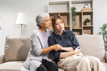 Fototapeta premium A mature mom and her young daughter, both Asian, sit on a sofa. The daughter appears sick and unwell, causing worry and possibly depression. The mom tries to encourage her and stay by her side.