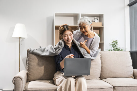 A mature mom and her young Asian daughter sit on a sofa, working with a laptop. The mom supports, encourages, and stays by her side, helping with warmth and caring love on Mother's Day.