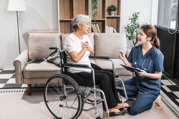 A young female nurse of Asian descent assists an elderly woman in a wheelchair, sitting on a sofa.