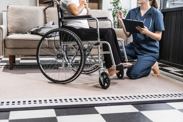 A young female nurse of Asian descent assists an elderly woman in a wheelchair, sitting on a sofa.