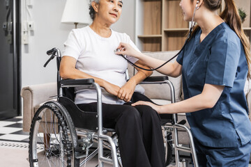 Fototapeta premium A young female nurse of Asian descent assists an elderly woman in a wheelchair, sitting on a sofa.