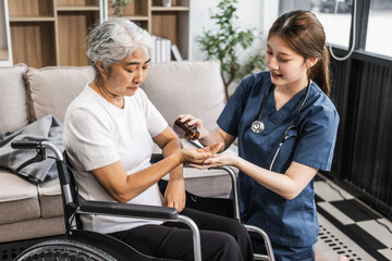A young female nurse of Asian descent assists an elderly woman in a wheelchair, sitting on a sofa.
