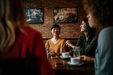Four Young Female Friends Meeting Sit At a Table In a Coffee Shop And Talk. Focus On A Asian Woman Talking