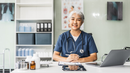 A mature female healthcare professional, confidently seated at her office desk in a hospital clinic, smiles as she works on her laptop, providing expert care to patients.