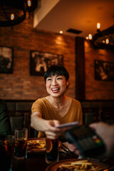 Happy mid aged Japanese woman paying bill with a contactless credit card in a restaurant