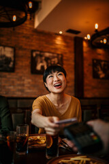 Happy mid aged Japanese woman paying bill with a contactless credit card in a restaurant