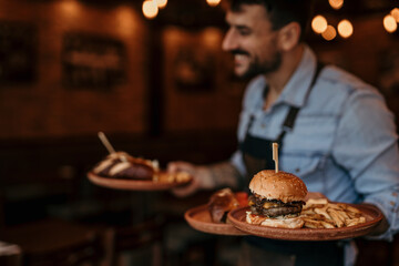 Smiling waiter in the restaurant serving two plates of burgers. Focus on a burger on plate