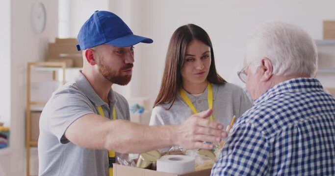 Volunteers team working at charity center giving free food donations of foodstuffs for poor mature people in charitable foundation. Humanitarian aid and social help concept. 4k video.