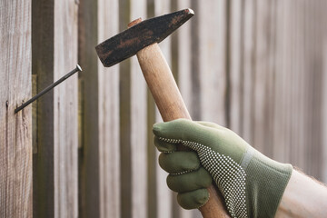 Man hands in gloves drive nail with a hammer in wooden rustic fence, carpentry.