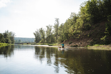 Obraz premium A person is peacefully paddling down a calm river, surrounded by a thriving forest landscape. The serene scene showcases water, sky, plant life, and the beauty of nature