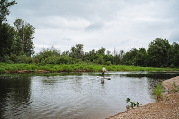 Fototapeta premium In the midst of a tranquil river, a person expertly balances on a paddle board, surrounded by trees, water, and a peaceful sky above, creating a serene natural landscape