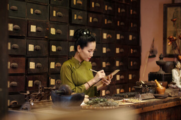 Asian young apothecary making list of herbs and spices in traditional Asian drug store