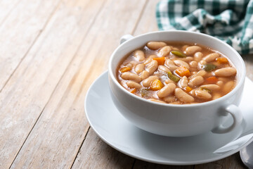 White beans soup with vegetables in white bowl on wooden table. Copy space