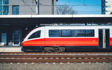 Fototapeta premium High-speed red passenger train at railway station platform under clear blue sky at sunset. Train station. Modern railway transportation concept. Railroad. Commercial. Urban rail transport in Austria