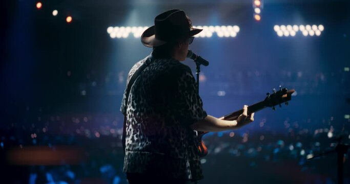 Country Singer Wearing Cowboy Hat Singing for the Audience while Playing Guitar. Performer Strums Acoustic Riffs, Singing Passionately on Stage. Concert Crowd is Immersed in the Performance