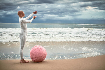 Full length portrait of hairless girl with alopecia in white futuristic suit standing on sea beach...