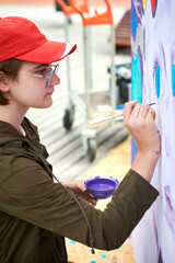 Female painter in red cap draws surreal picture with paintbrush on canvas for outdoor street...
