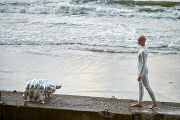 Young hairless girl with alopecia in white futuristic suit walking on concrete fence with toy...