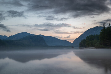 Fototapeta premium A picturesque view of Almsee Lake with Wooden Hut in Austria at sunset. The sky is filled with beautiful, colorful clouds, and the mountains in the distance are reflected in the calm water. 